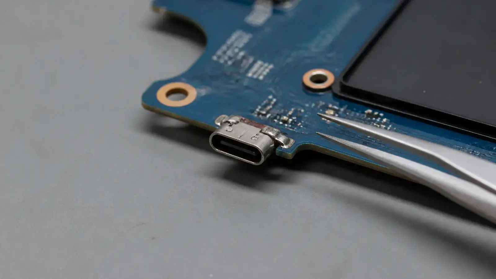 Крупно край платы ноутбука с разъёмом USB-C, который немного отошёл от платы, рядом пинцет