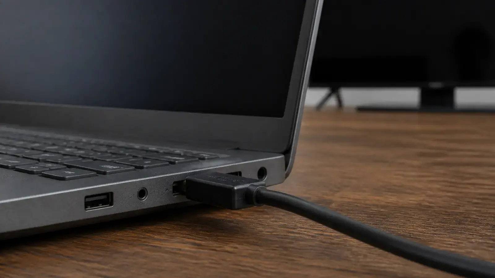Крупно боковая грань ноутбука с портом HDMI, в который неровно вставлен чёрный кабель, на размытом фоне телевизор