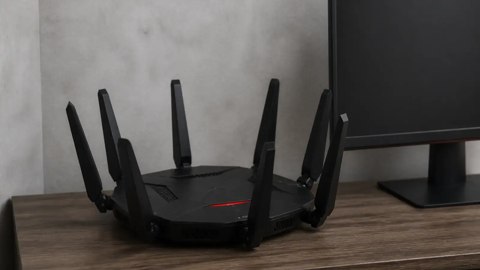 Чёрный игровой Wi-Fi роутер с восемью внешними антеннами и красной полосой подсветки стоит на деревянной тумбе рядом с тёмным монитором