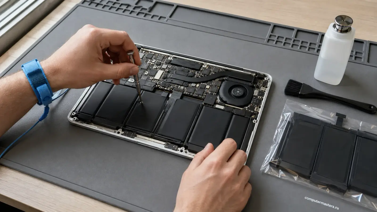 MacBook со снятой нижней крышкой на антистатическом мате: видны плата, вентилятор и батарея, мастер в браслете держит отвёртку, рядом новая батарея в упаковке, кисть и спирт