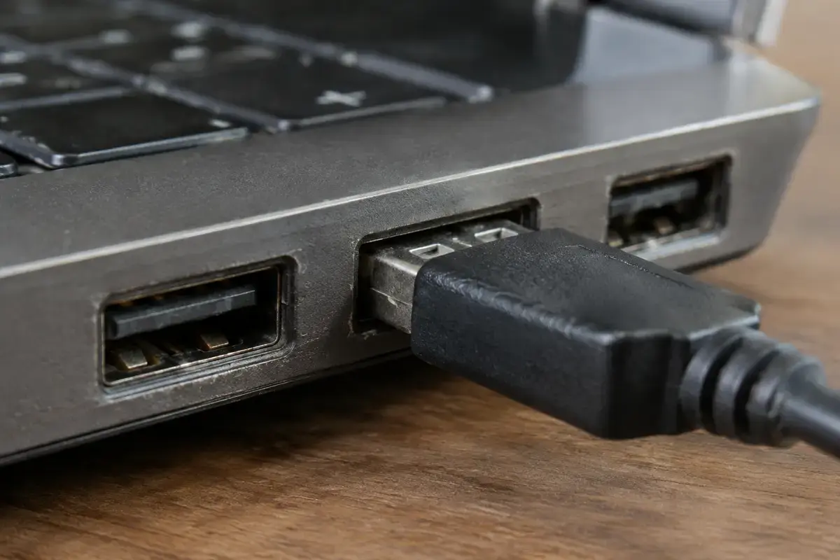 Крупный план боковой стороны ноутбука с тремя портами USB-A, в среднем порту чёрный штекер, металл вокруг портов потёртый