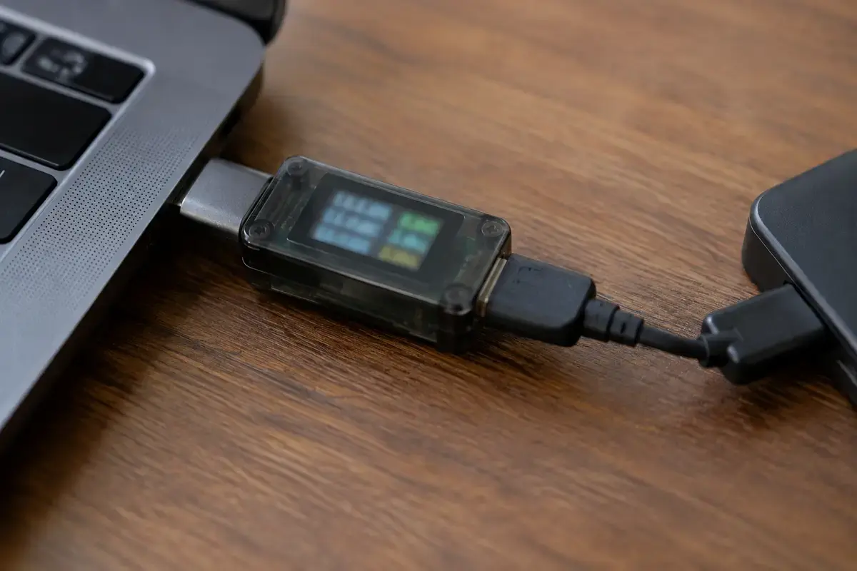 Прозрачный USB-тестер с цветным дисплеем включён между портом ноутбука и чёрным кабелем внешнего диска