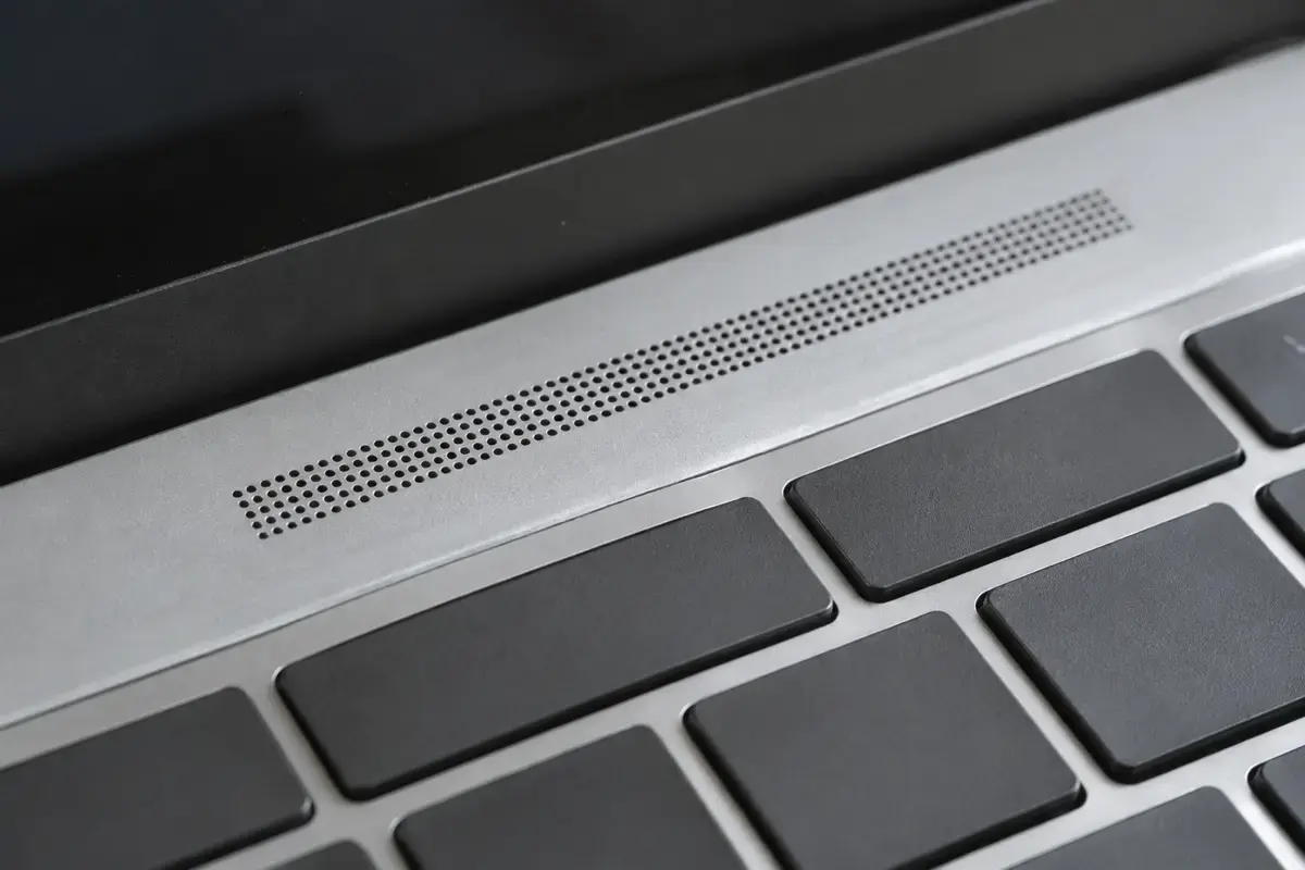 Крупный план узкой решётки динамика из мелких отверстий над клавиатурой серебристого ноутбука, рядом тёмные клавиши